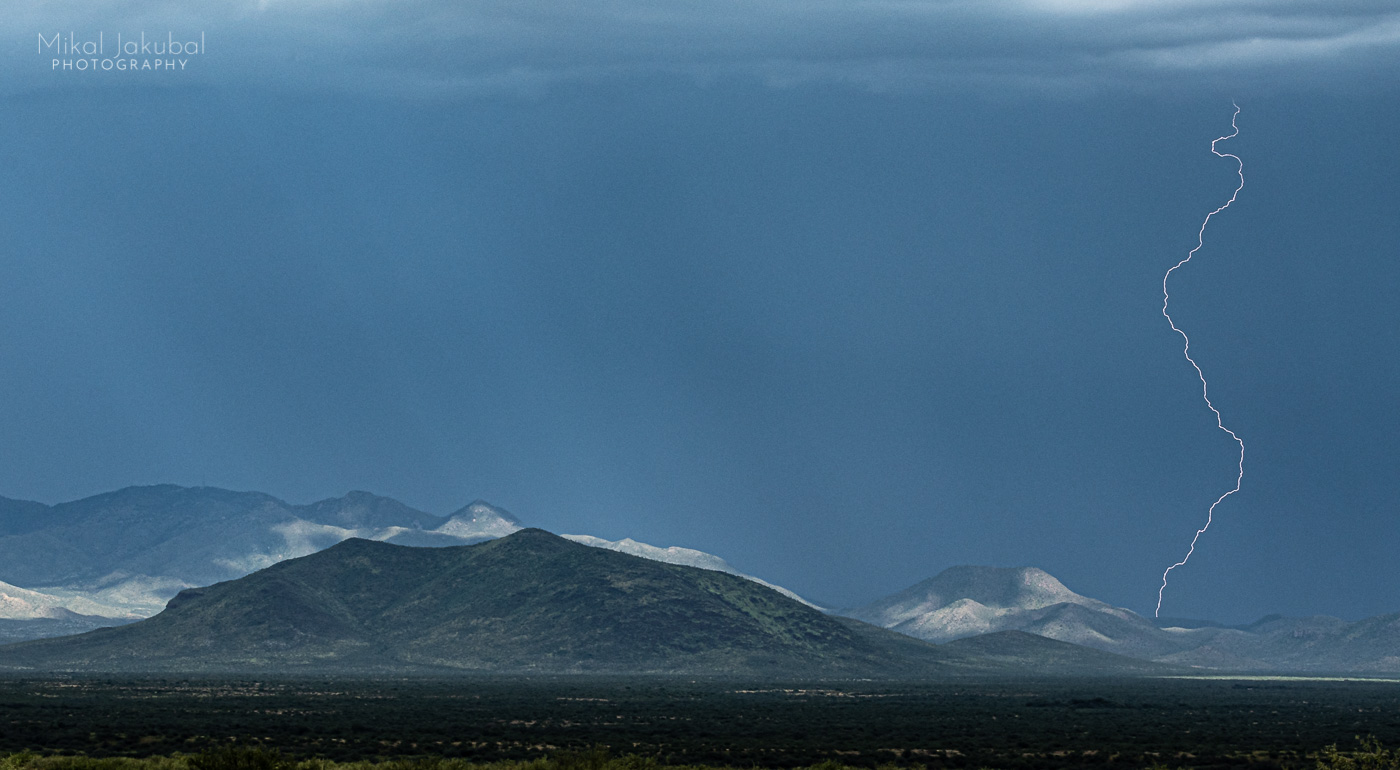 A deep blue gray sky is cut by a thin thread of lightning in the distance. To the left are rounded, shadowed hills.