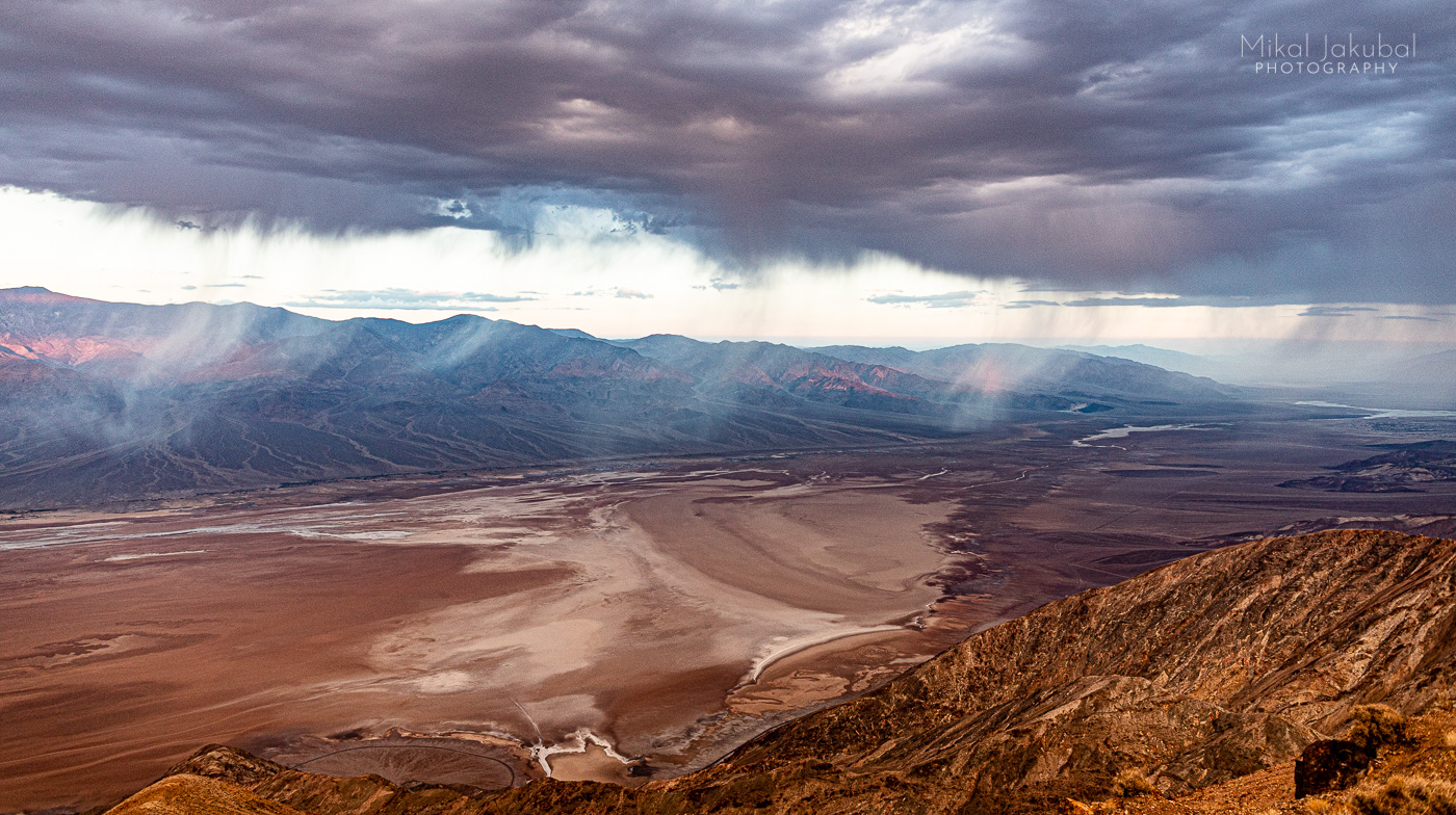 Looking out over Death Valley, with high storm clouds, undersides lit up by dawn light. Below the rocky landscape sloped down to brownish white salt flats.