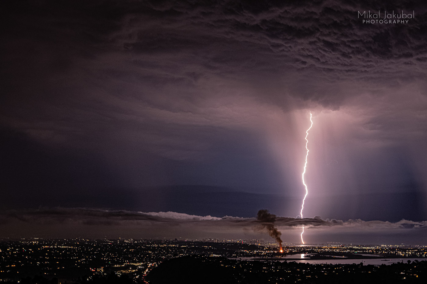 The scene is nighttime over the city of San Diego. A purple bolt of lightning drops straight down from purplish rain clouds. Almost in line with the bolt, a large flame shoots up and a smoke cloud drifts off from burning palm trees that were hit by a previous strike.