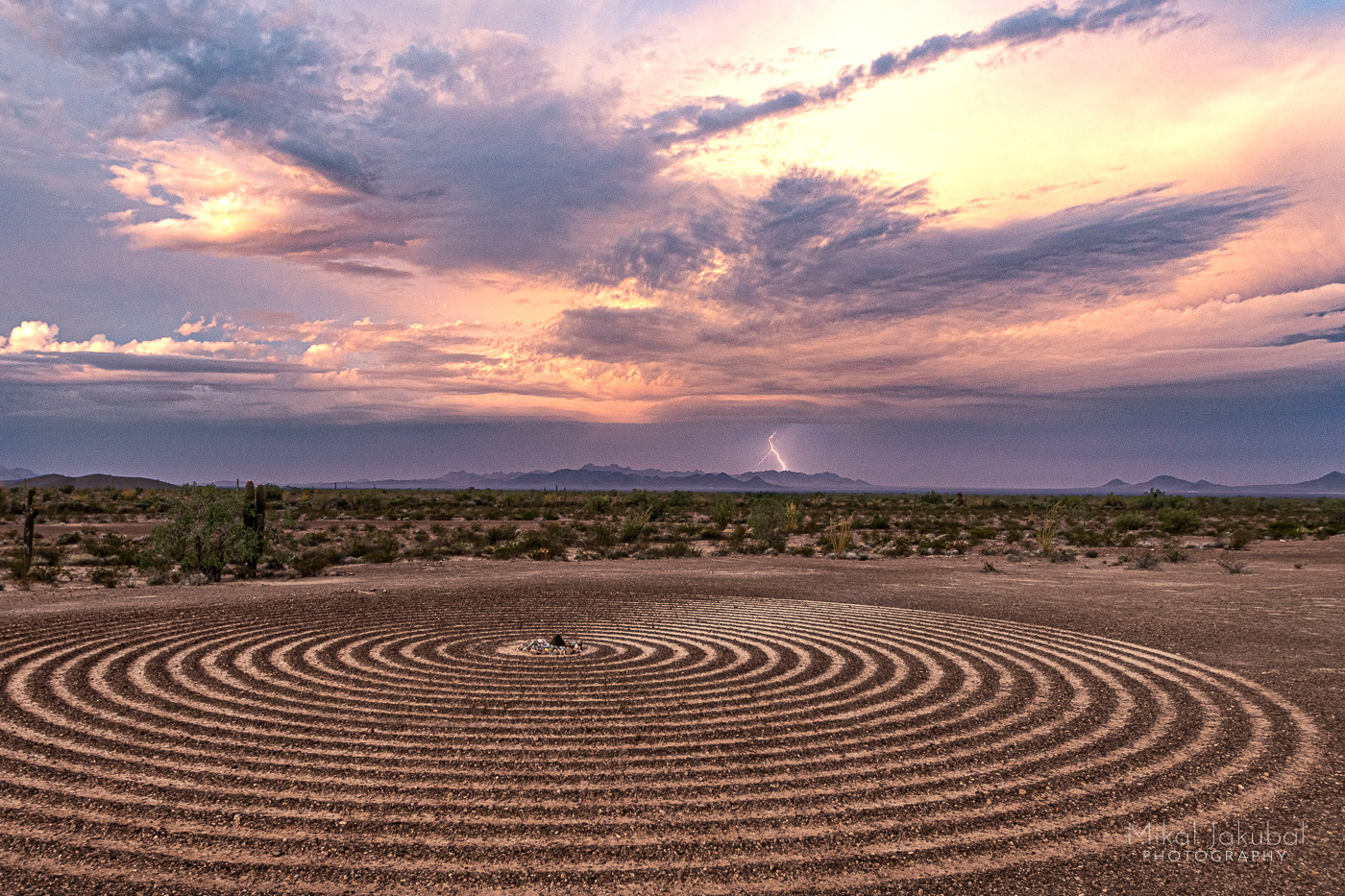 In the foreground, the Spiral Labyrinth forms a 60-foot diameter circle of tight concentric rings alternating between dark gravel piled into ridges and the lighter soil exposed beneath. In the background, bright orange and purple sunrise clouds are punctuated by a single bolt of lightning on the far horizon.