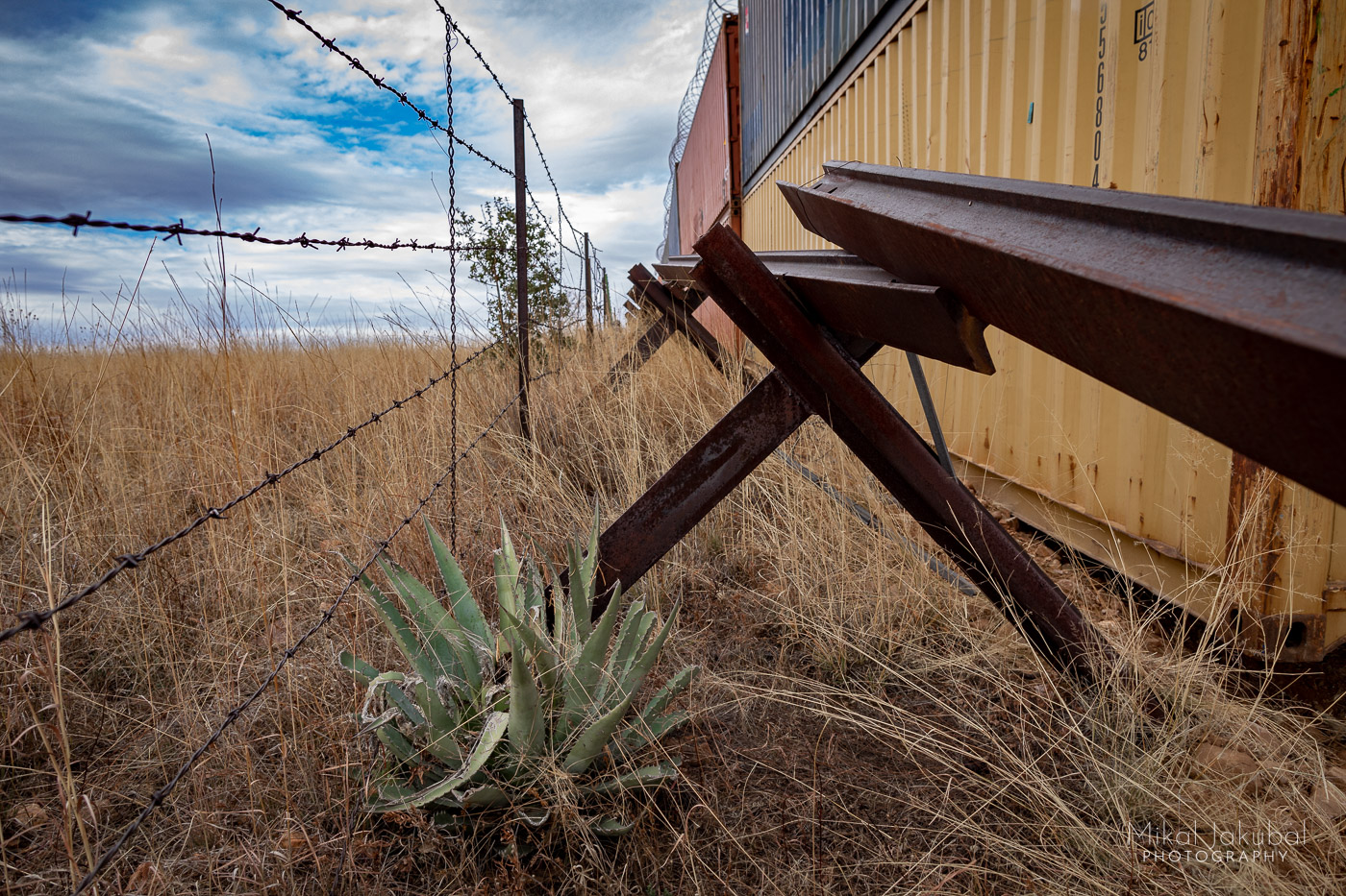 Photo shows a green agave plant in the foreground. Three types of fence — a barbed wire fence; a vehicle barrier made from welded pieces of railroad track; and the container wall made from double-stacked shipping containers — form three parallel lines into the distance along the U.S.-Mexico border.