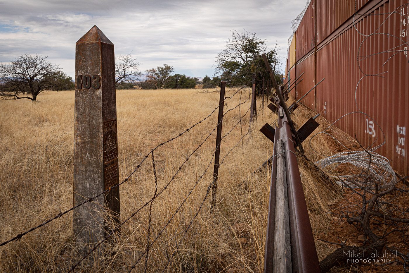 Looking into the distance along the border wall. To the far left, a concrete obelisk with the number "103" stands in the grass. Next to it is a barbed wire cattle fence. Center frame is a 3-foot tall barrier made from welded railroad rails. To the right is a line of double-stacked shipping containers. A shiny new roll of concertina wire loops down from the top of the containers.