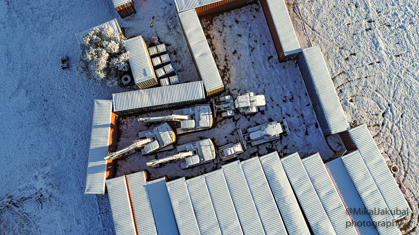 Aerial view of snow covered shipping containers arranged like a walled city, with snow covered heavy equipment parked inside.