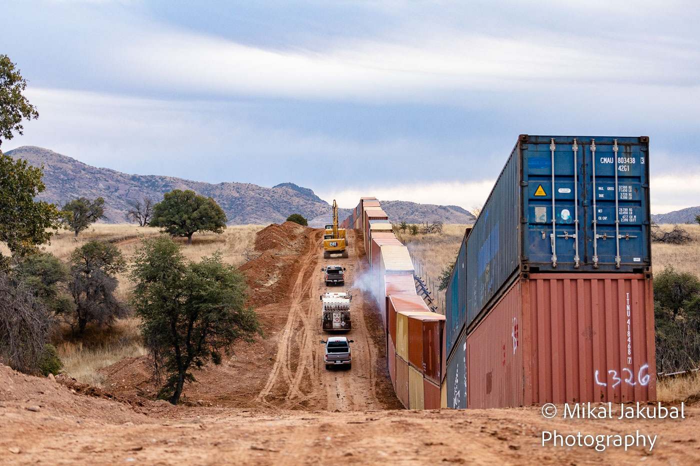A line of heavy equipment and pickup trucks drives away along a dirt road next to a double-stacked line of shipping containers.
