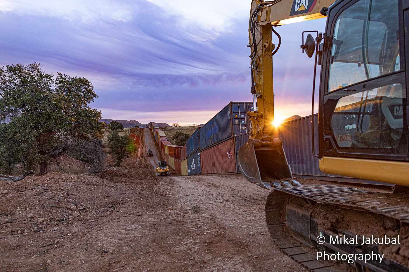 A large yellow excavator enters the photo from the right as the sun flares next to the bucket at dawn. Past the excavator, there is a line of double-stacked shipping containers next to a dirt road.