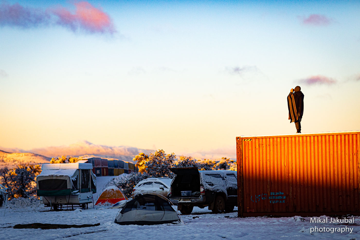 A protester with binoculars stands atop a sunlit shipping container at dawn. In the shadow below is a collection of snow covered tents and vehicles.