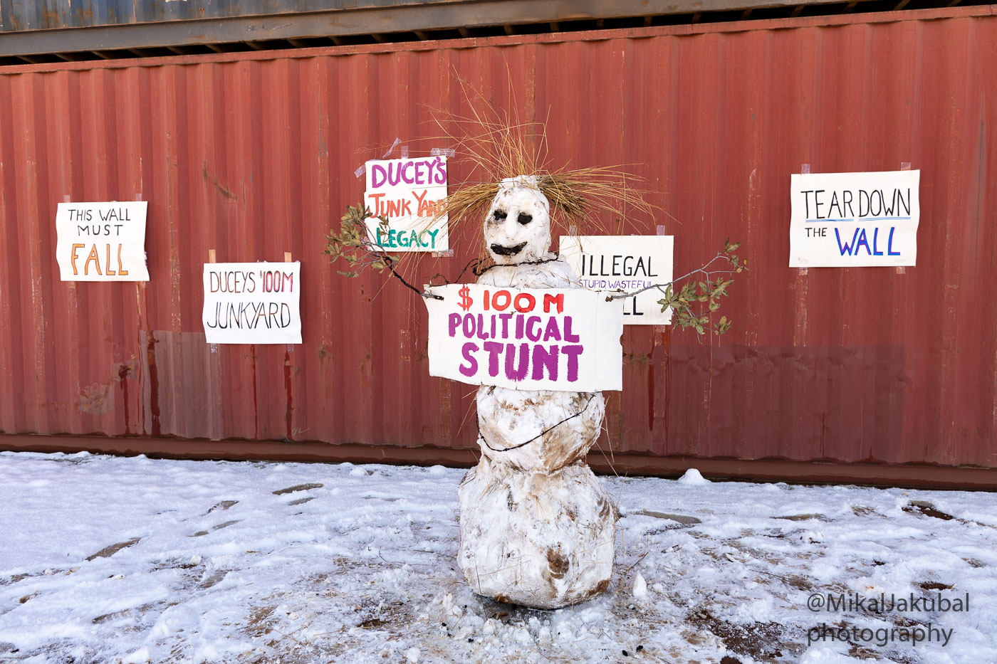 A dirty snowman made from found large snowballs and branches for arms holds a protest sign that reads "$100 million political stunt". Behind it is a shipping container with additional signs saying "Tear down the wall," "this wall must fall," " Ducey's junk yard legacy."