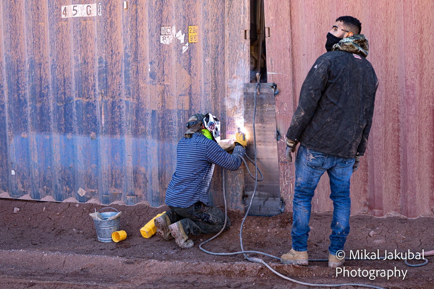 One worker in a welding helmet tack welds a steel plate to cover the gap between two shipping containers while a second worker looks away from the bright welding arc.