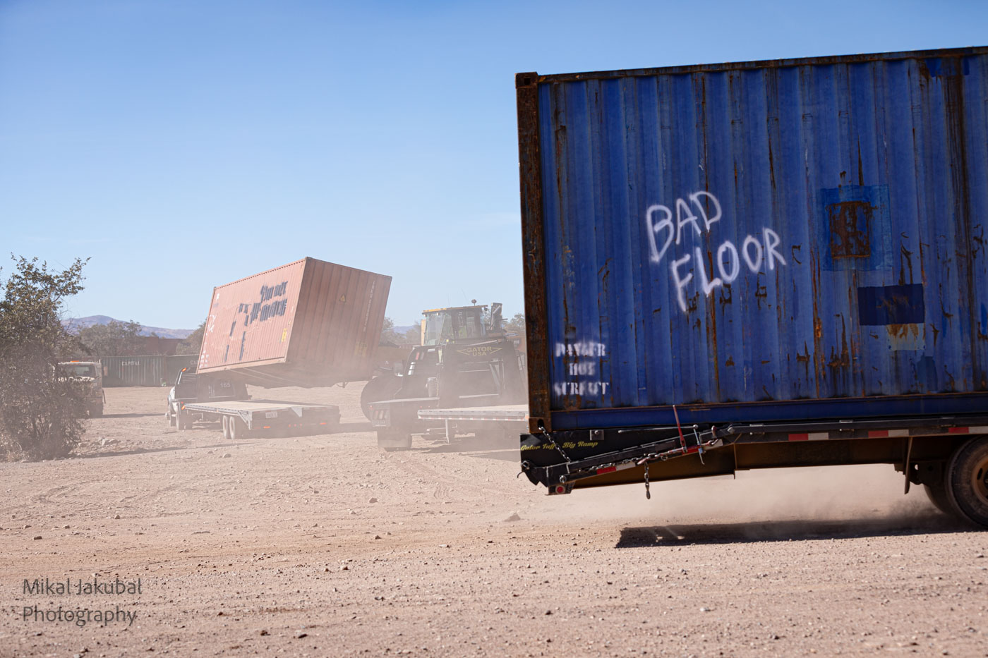 In the foreground, a blue shipping container sits on a trailer. It has the words "bad floor" spray painted on the side, a reference to why the container was rejected by shipping companies and sold as surplus. In the background a large forklift is kicking up dust as it loads a shipping container onto a flatbed trailer.