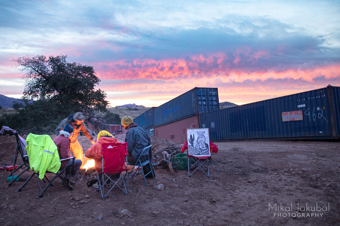 Four people gather in chairs around a small campfire at dawn, with magenta clouds to the east. Nearby in the shadows, shipping containers are stacked up to form a wall along the US-Mexico border.