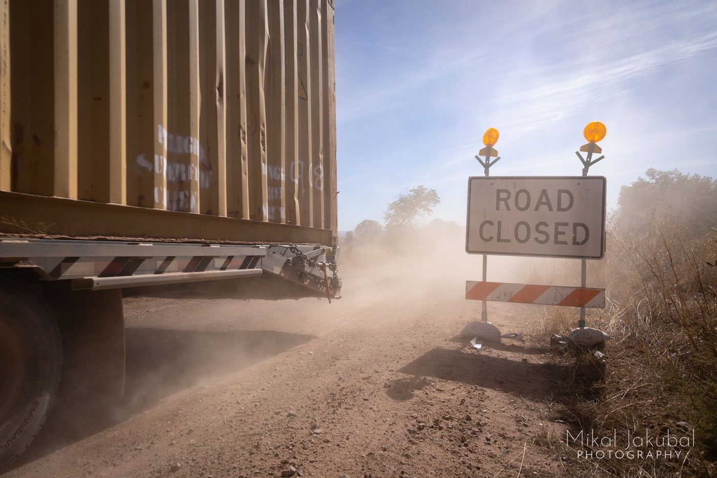 A dusty roadside scene. A "road closed" sign faces the camera while the last end of a shipping container on a trailer leaves the scene to the left, its wheels kicking up dust clouds.