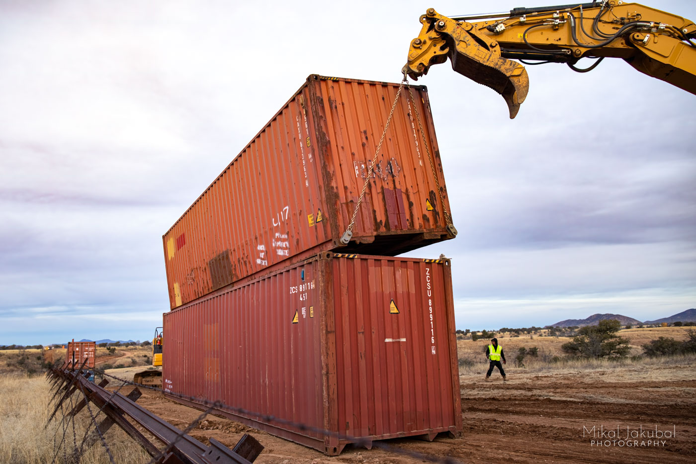 The steel arm of a large excavator lifts one shipping container off the top of another one with two large chains. The container appears to be about to tip over, but is in fact held securely by the chains. A worker in a yellow vest walks nearby.