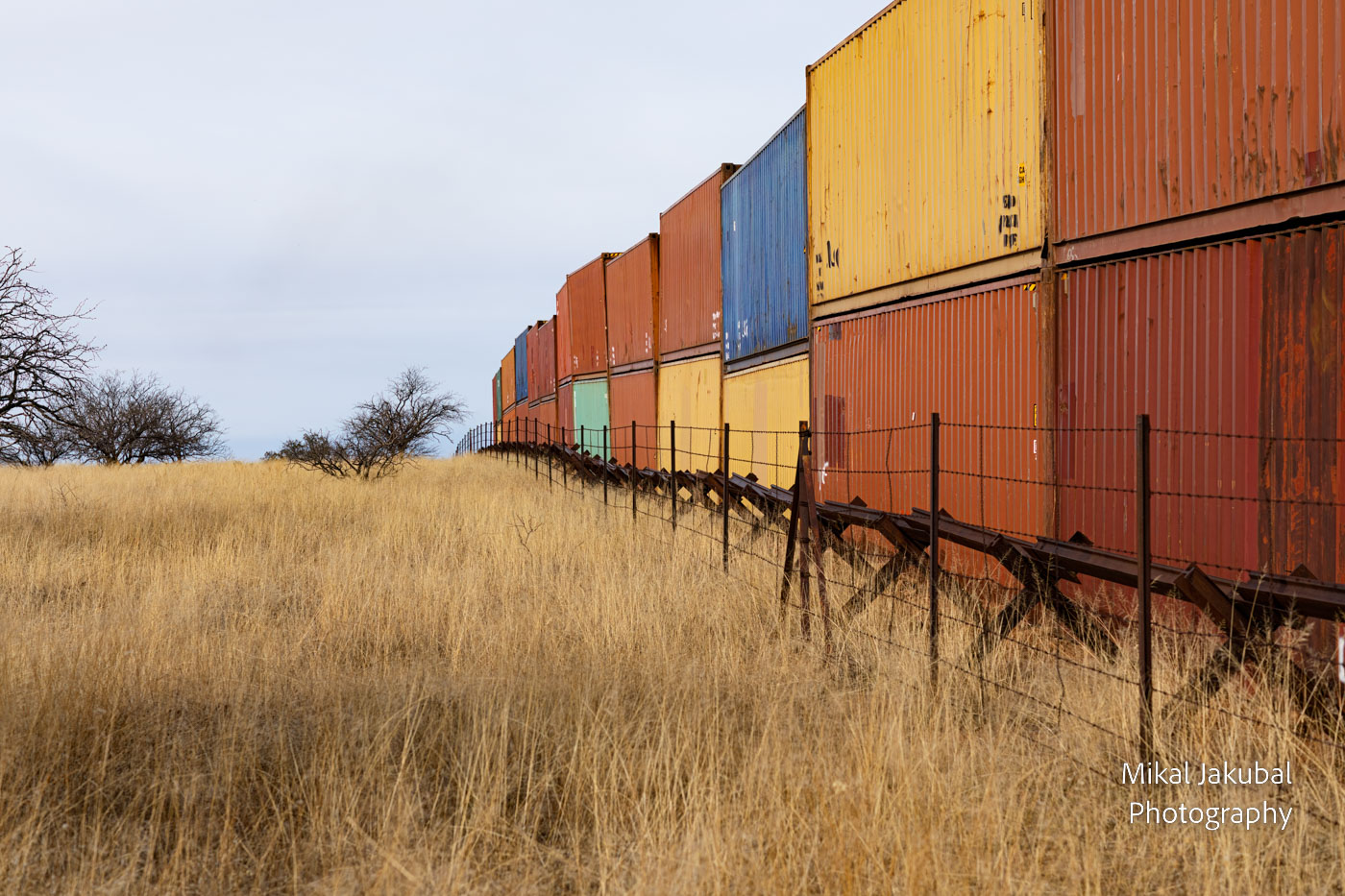 A line of double-stacked shipping containers forms a wall along the U.S.-Mexico border. Next to it is a low barrier made of welded railroad rails. Short chunks of rail are welded into X shapes and then horizontal pieces of rail are then laid across them and welded.
