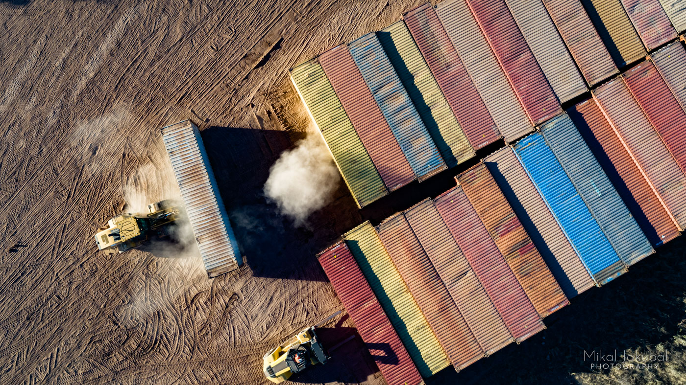 Two rows of multi-colored shipping containers are seen from above. At the end of each row is a large forklift. The one on the left has just picked up a container, stirring up a large dust cloud.