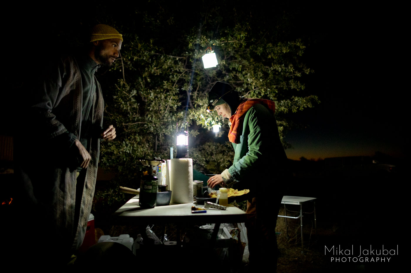 Two people, a man and a woman stand next to a small table in front of a tree at night. The table is illuminated with several small battery powered LED lights. The woman is preparing food on a small portable cook stove.