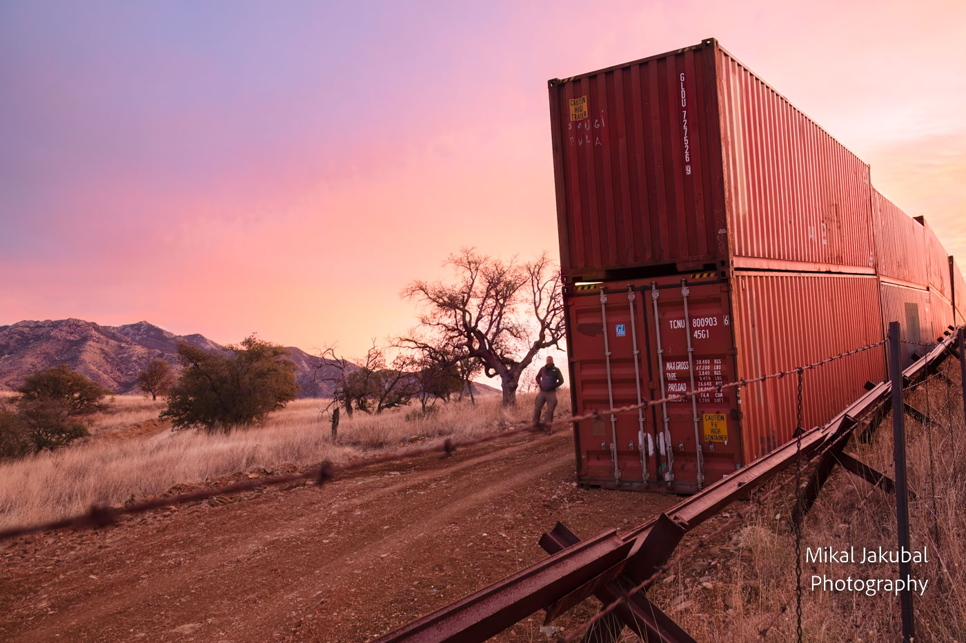 At dawn the sky is red, pink and purple. To the left in the background is a grassland with oak trees and mountains in the distance. In the center of the frame is a line of double-stacked red shipping containers. To the right of that is a barbed wire fence and a vehicle barrier made of welded railroad rails. At the left edge of the containers is a shadowed human figure against the skyline, hands in pockets looking at the camera.