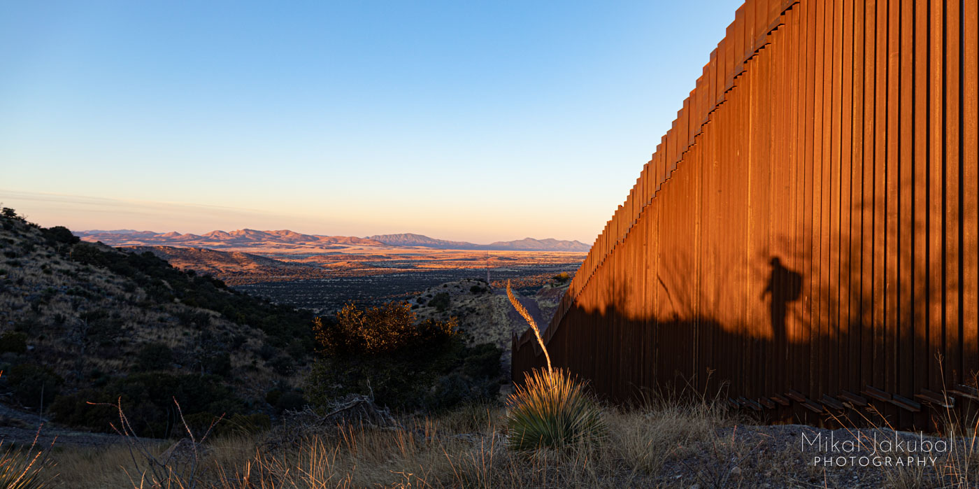 A photo of the U.S.-Mexico border area. The 30' tall border fence, consisting of rusty-orange square steel tubes with gaps fills the frame to the right. The rest of the frame shows the vast landscape along the border in the distance. A shadow of a person wearing a backpack is cast against the wall, but the person is not visible in the photograph.