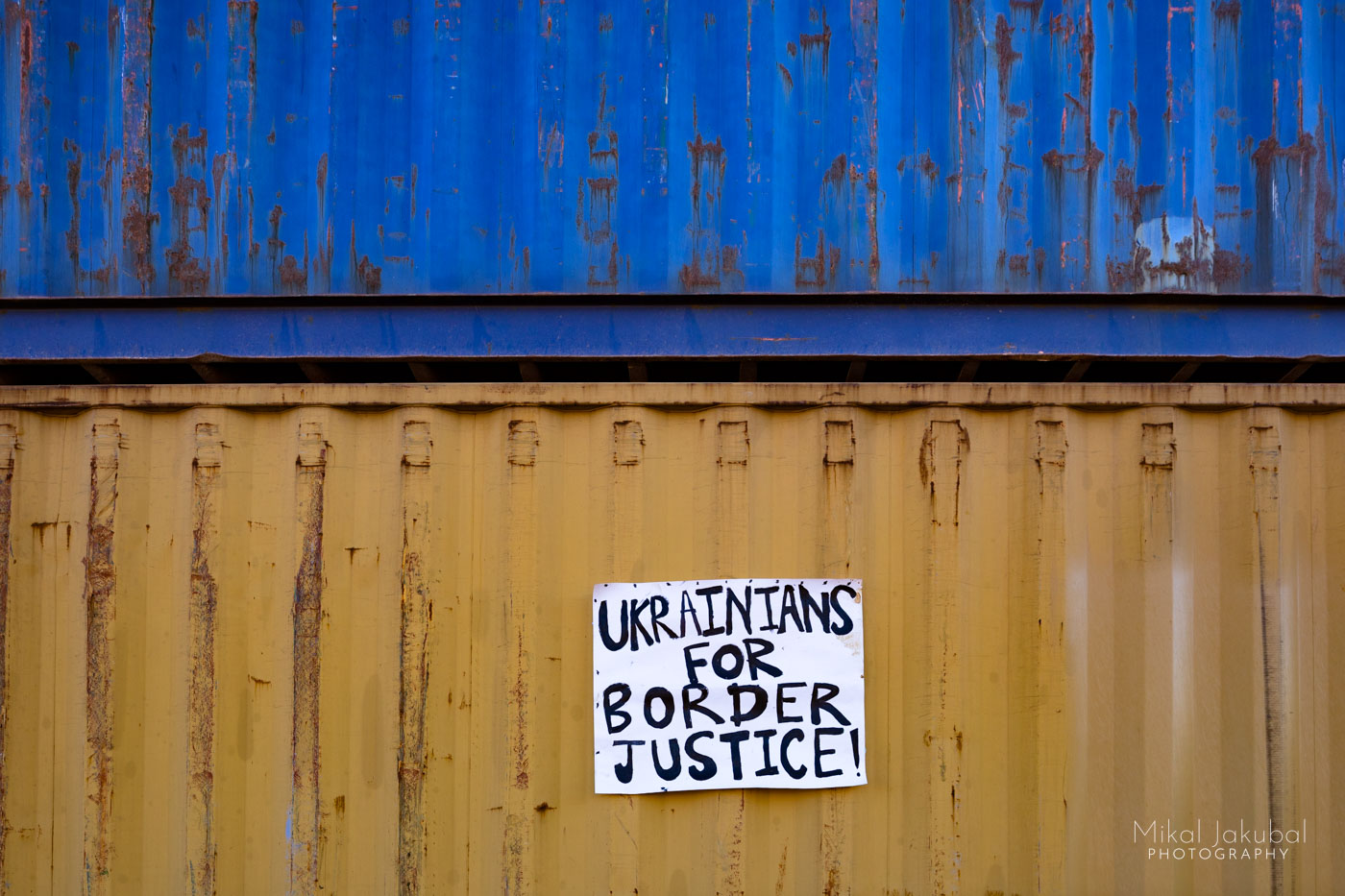 A close up view of a blue shipping container stacked on top of a yellow one, mimicking the colors of the Ukrainian flag. On the yellow container is a hand-lettered sign that says "Ukrainians for border justice!"