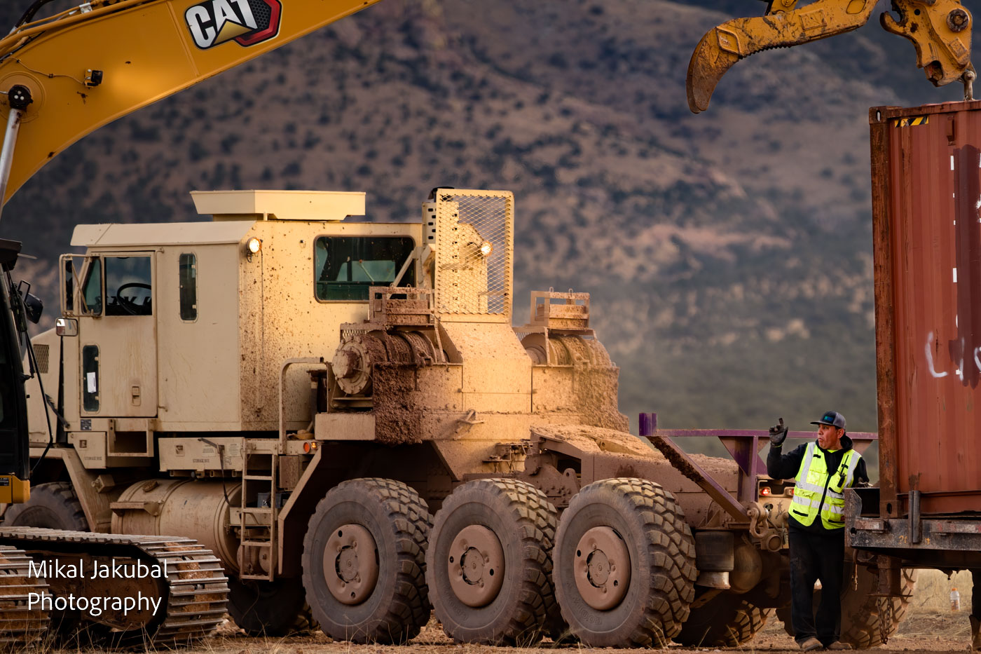 A worker in a high-vis vest gives a hand signal to the operator of a large excavator that is lowering a shipping container onto a giant flatbed trailer. The trailer is towed by an 8-wheel, yellow military tractor vehicle.