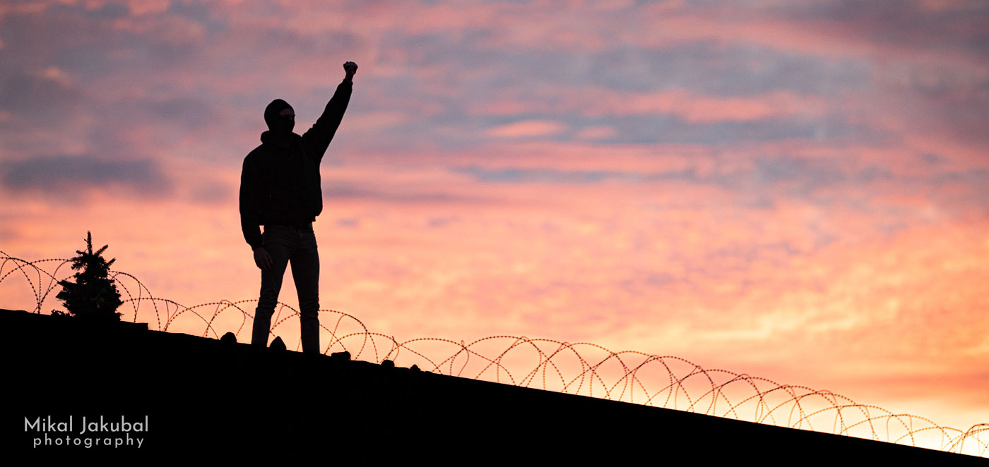 A person stands silhouetted with fist in the air atop a shipping container topped with razor wire. The sky is sunset pinks, yellows and oranges.