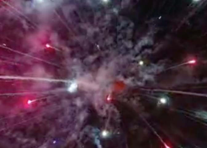 A dark sky with fireworks exploding close up, giving off smoke and sending green and red streamers out from the center of the explosion.