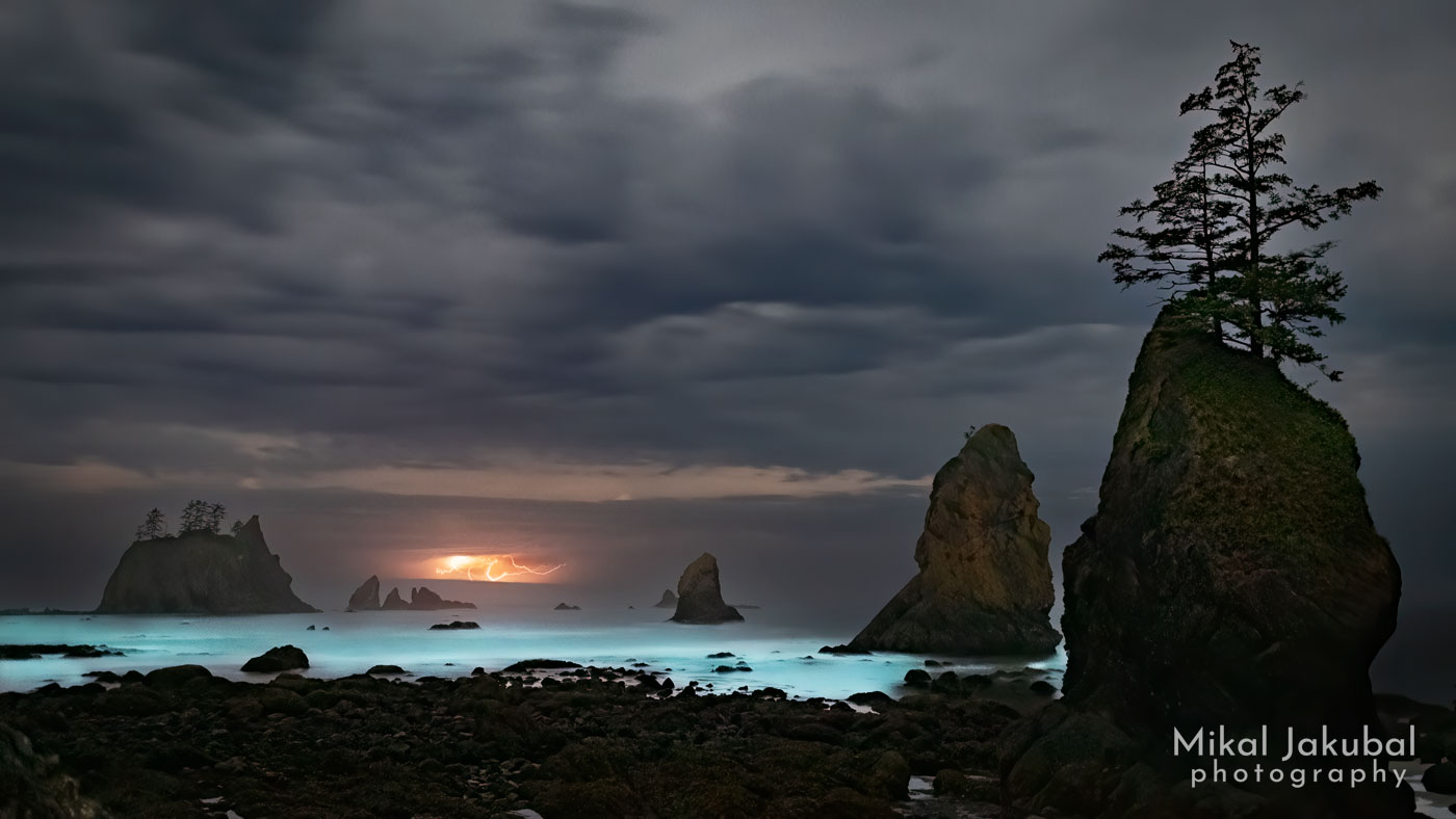 A rocky shoreline at night under storm clouds. A small orange bolt of lightning strikes the ocean in the far distance. Near shore, a ghostly blue glow from bioluminescent plankton lights up the water.