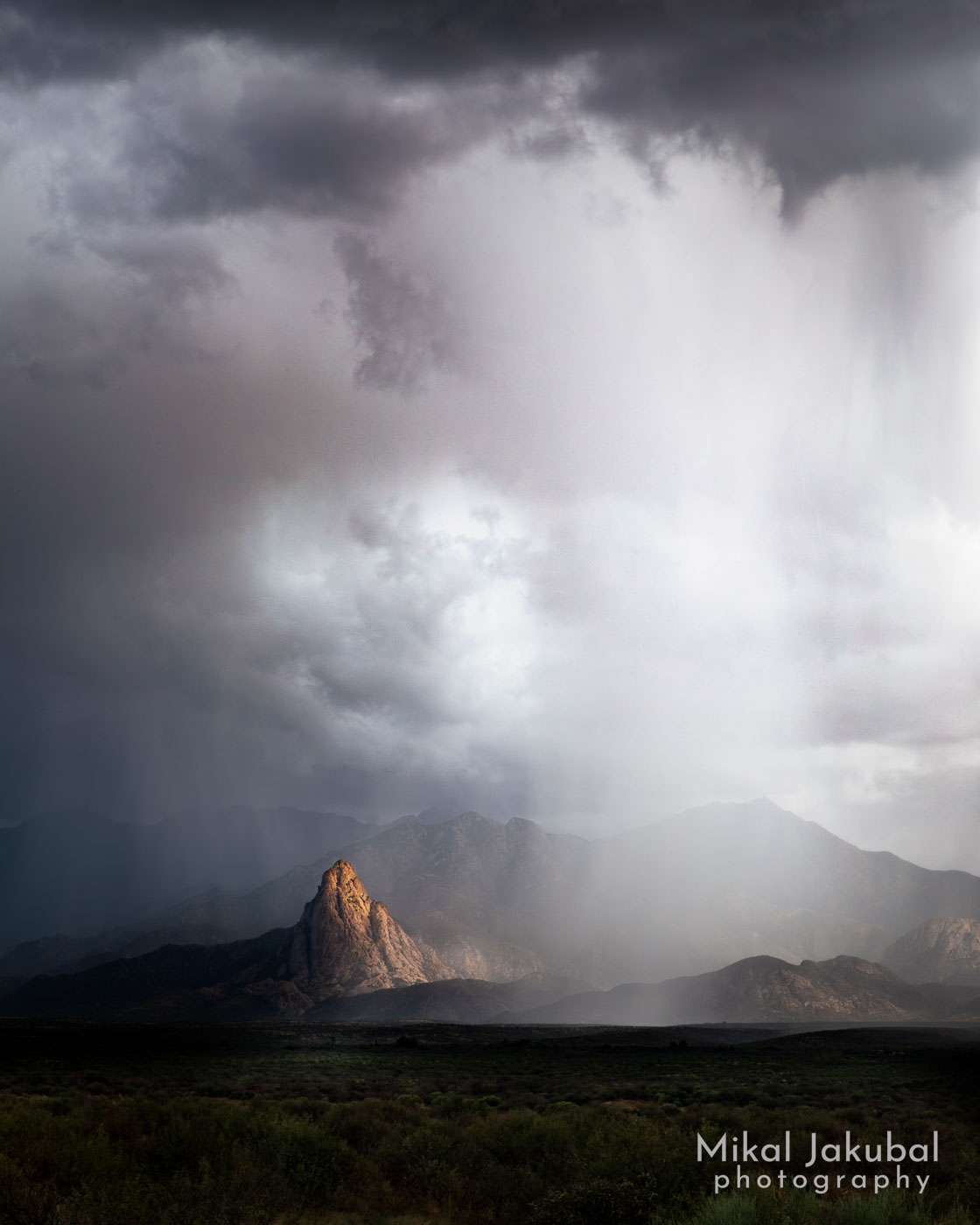 A stormy day, with rain falling from high, dark clouds. Rain shrouded mountains lie behind the clouds, but in the foothills a rock outcrop is illuminated by sunlight through a cloud break.