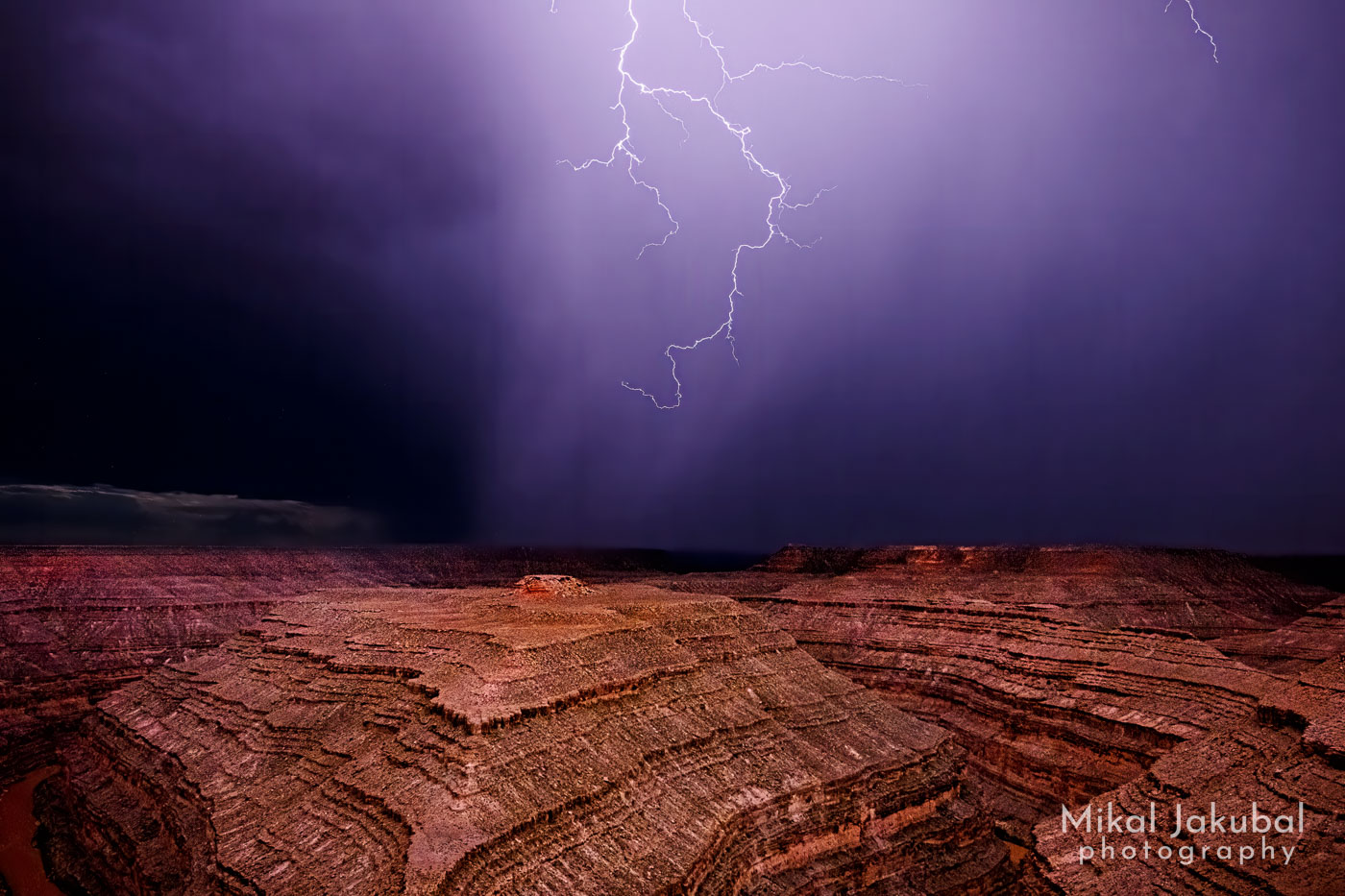 A bold of lightning descends through purple clouds at night, illuminating a layered bedrock canyon below.