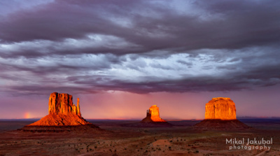 Desert towers rising from a broad plain are illuminated by bright orange alpenglow under a dark, foreboding sky at sunset.