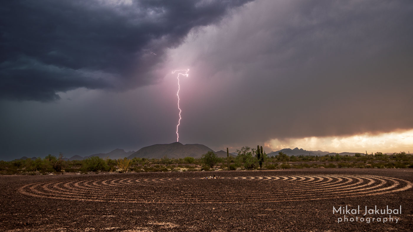 A purple tinged lightning bolt descends from dark storm clouds, striking a small hill in the desert. In the foreground is a 60' diameter spiral labyrinth scraped into the stony desert soil.
