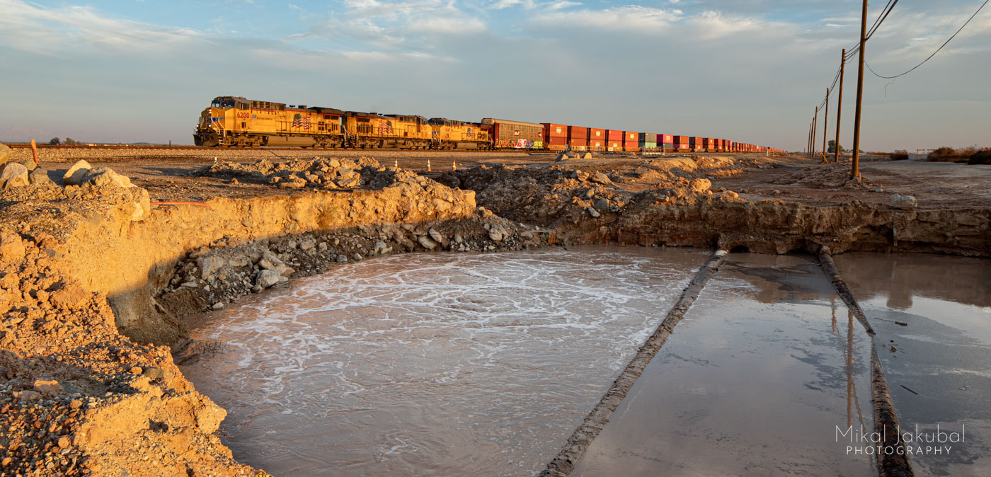 The Niland mud spring, also known as the Mundo mud spring and the Niland geyser just before sunset. The spring is an 80' wide hole of muddy water with a bubbling gas upwelling at the left side. Two old pipes cross at the water's surface and fiber optic cables are strung on poles overhead. In the background, a long freight train is about the leave the frame and extends into the distance. The three engines are yellow, followed by lines of mostly orange colored freight containers on flatbed cars.