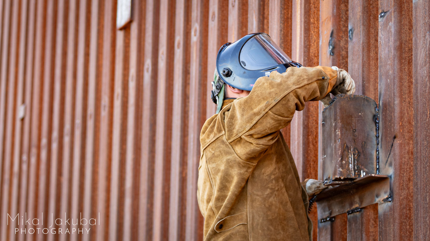 A worker in a welder's helmet and a heavy brown leather welder's coat places a grapefruit size rock into a steel canister that is welded onto the steel bollards of the US-Mexico border fence.