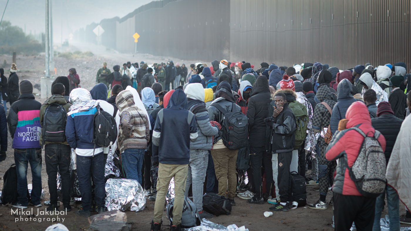 The U.S.-Mexico border fence looms in the background and recedes into the mist and distance just after dawn as a group of a couple hundred people stands around awaiting direction from Border Patrol.