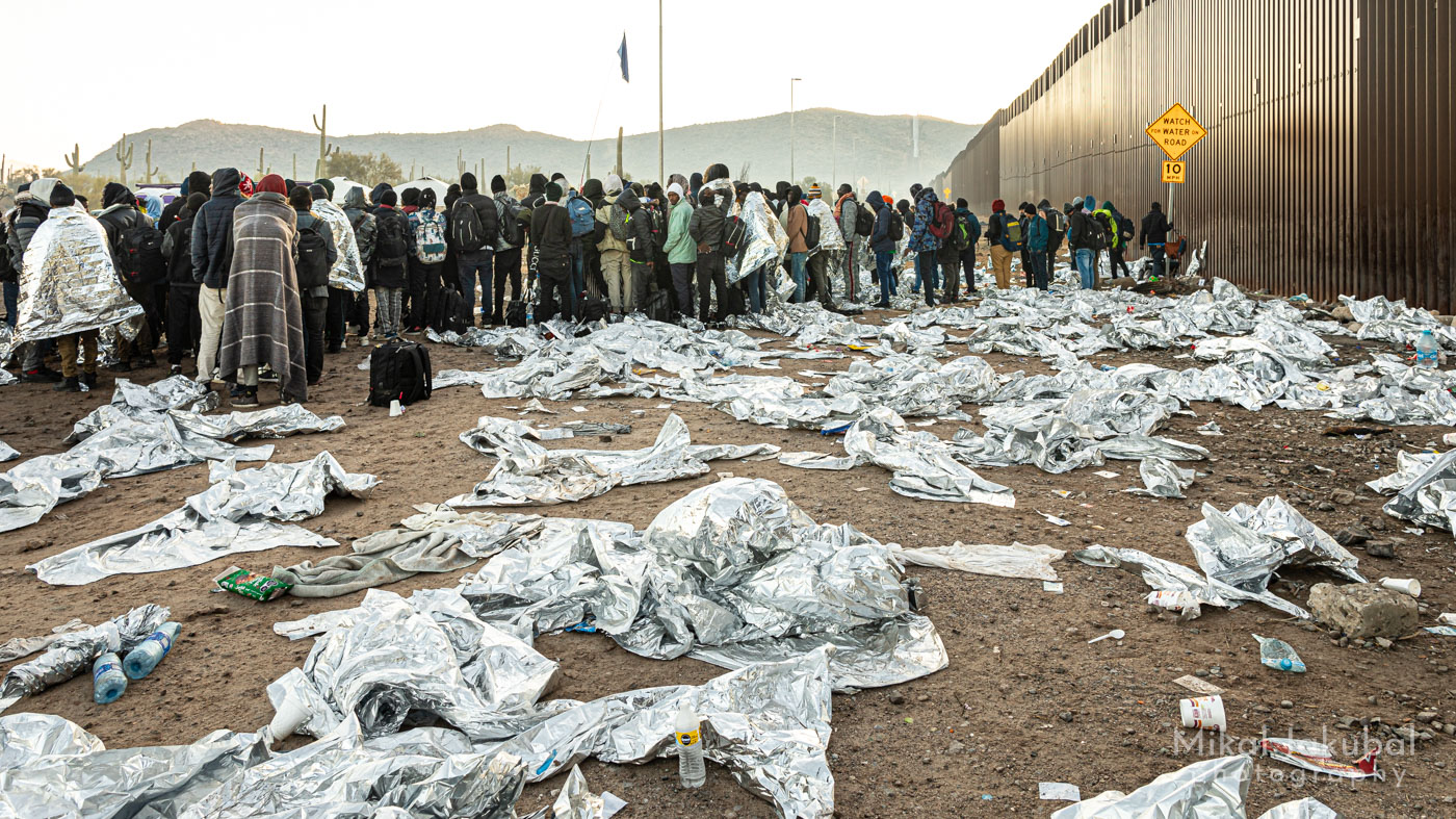 A large group of people, seen from behind, lines up near the 30' tall U.S.-Mexico border fence at dawn. In the foreground, hundreds of crumpled mylar "space blankets" cover the ground.
