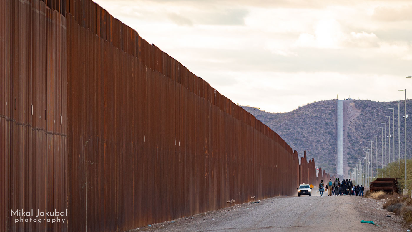 The U.S.-Mexico border fence, built of rows of 30' tall, 6" x 6" square steel tubing set into concrete 4" apart, seen in a compressed telephoto image looking down the parallel road. The fence cuts a straight line across the landscape and up a distant hill. In the mid-ground, a cluster of about 40 people walks away as a white Border Patrol truck drives toward the camera.
