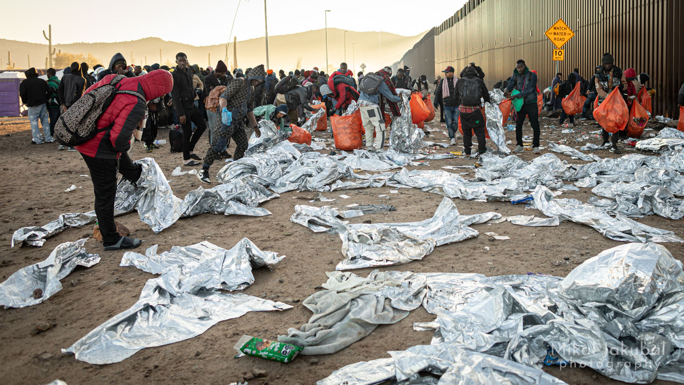 To the right is the 30' tall U.S.-Mexico border fence. In front, the ground is covered with discarded mylar "space blankets" used by people for warmth during the night. Asylum seekers were given orange plastic trash bags by Border Patrol and are now picking up the blankets and other trash.