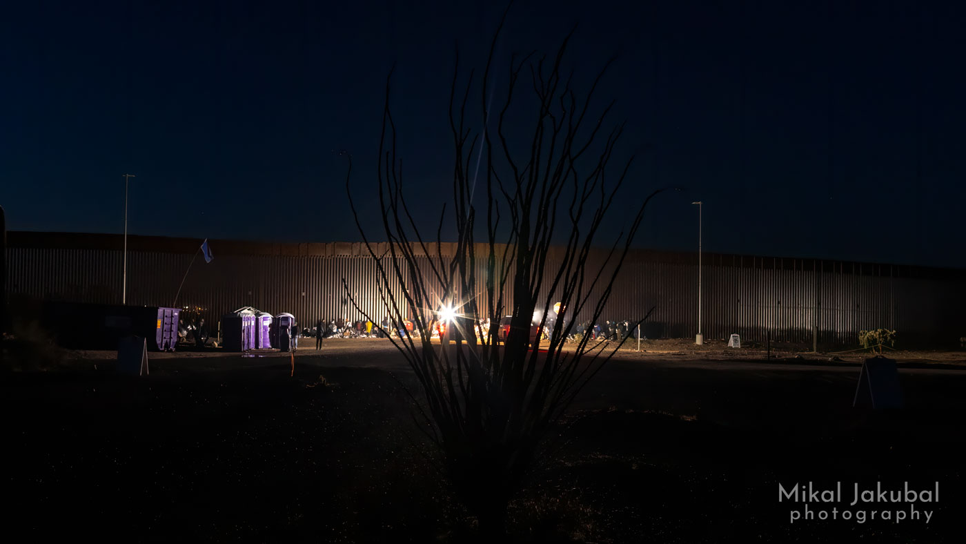 A nighttime seen along the U.S.-Mexico border fence. In the background there is a line of migrants sitting and standing near the fence, illuminated by the headlights of a Border Patrol pickup truck. To the left is a line of purple port-a-potties. Silhouetted in the foreground is a leafless ocotillo bush, it's upward-pointing branches forming a vase shape against the background.