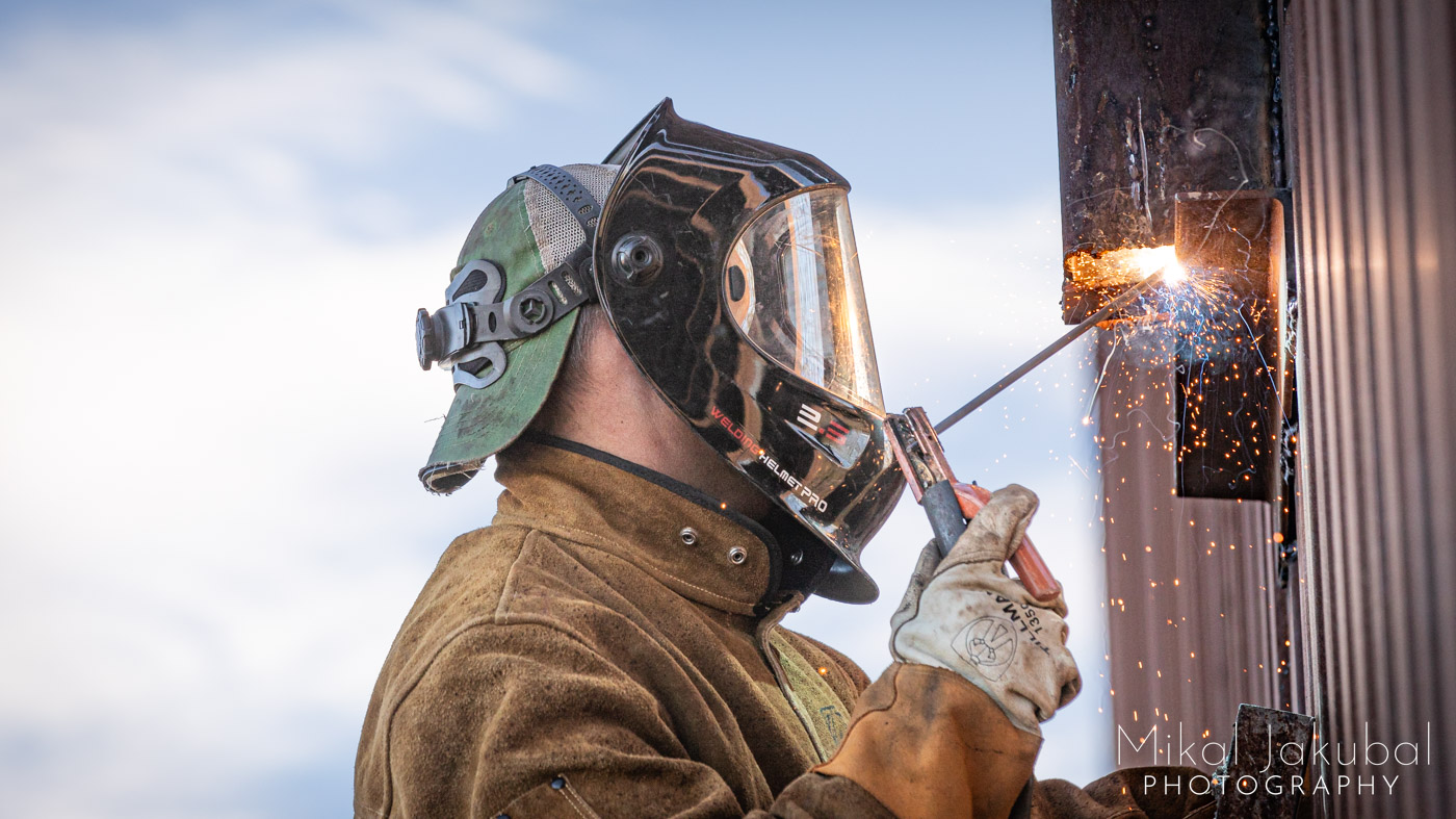Close-up of a welder in a welding helmet and thick leather welding jacket attaching a piece of steel to the US – Mexico border fence. Little red sparks are flying everywhere and the welder's work and face are illuminated by the bright white of the arc.