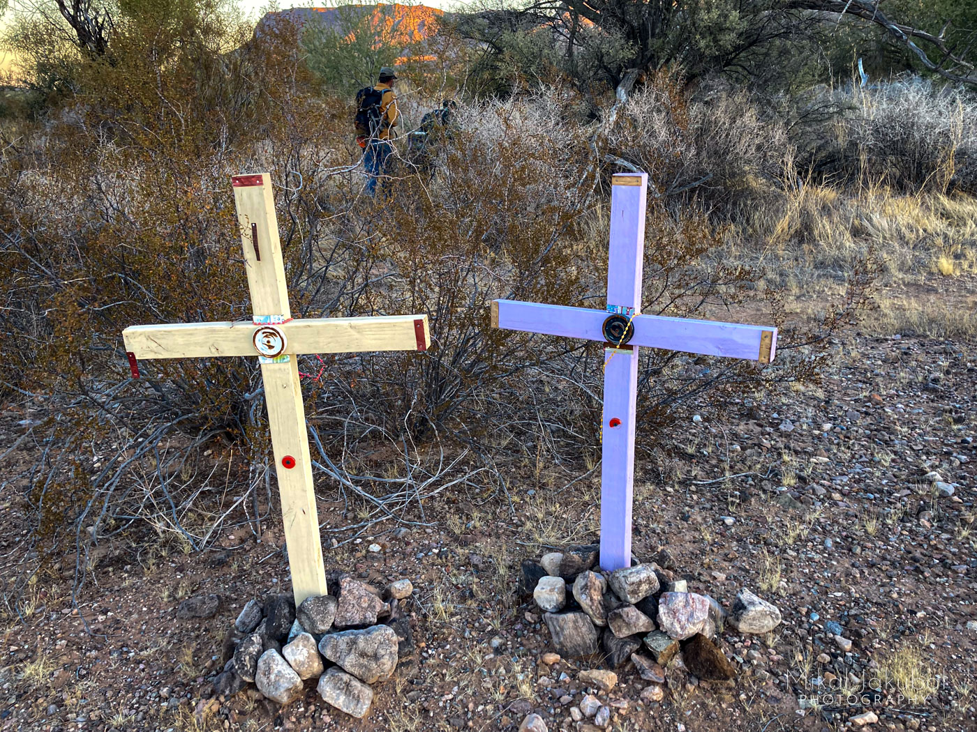 Two crosses, one yellow and one lavender, each about 3' tall and propped up with small piles of rocks are seen surrounded by desert scrub bushes. In the background, two hikers make their way through the brush.
