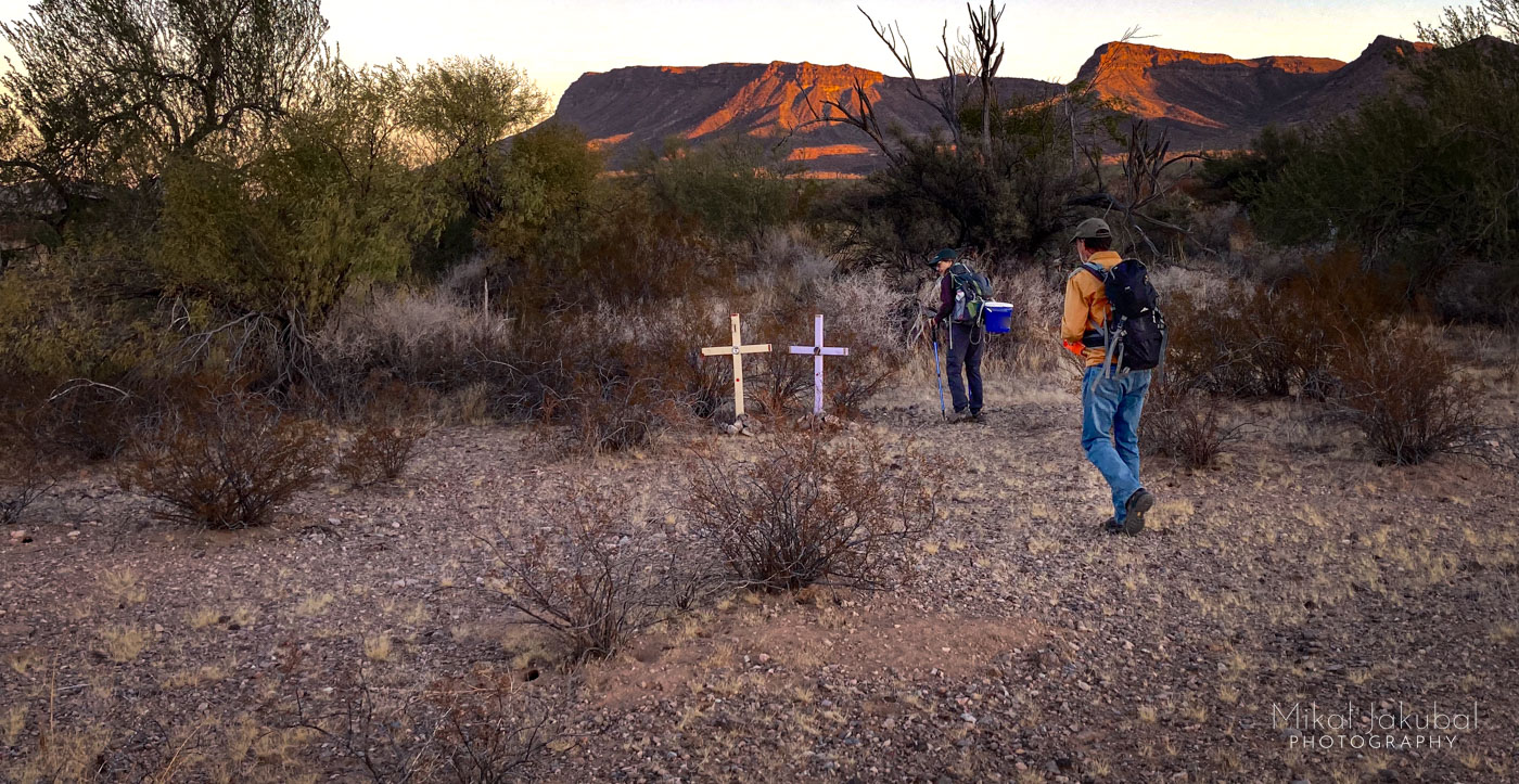 Two hikers are seen walking through coarse desert shrubs with first light hitting a mountain range in the background. To the hikers' left are two crosses, one pale yellow, one lavender, marking the spot where two dead people were found.