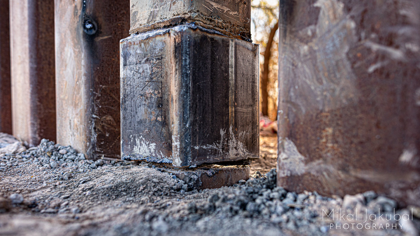 A close-up view from ground level of the base of the US – Mexico border fence. One of the 6" x 6" steel posts has been torch-cut entirely through, leaving a quarter inch gap.
